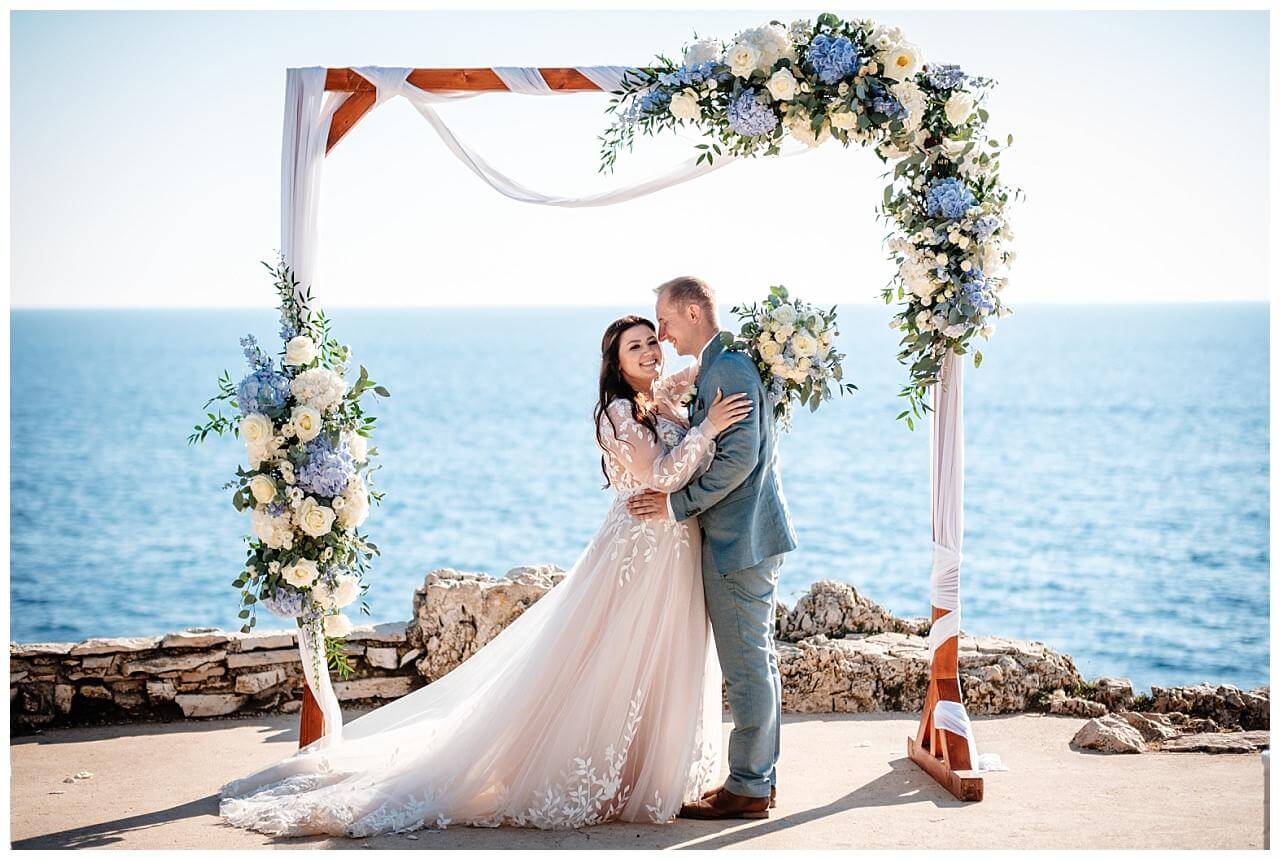Freie Trauung mit dem Blick aufs Meer und Traubogen aus Holz mit weißen Stoff und blau weißen Blumen bei einer Hochzeit in einer privaten Finka in Kroatien