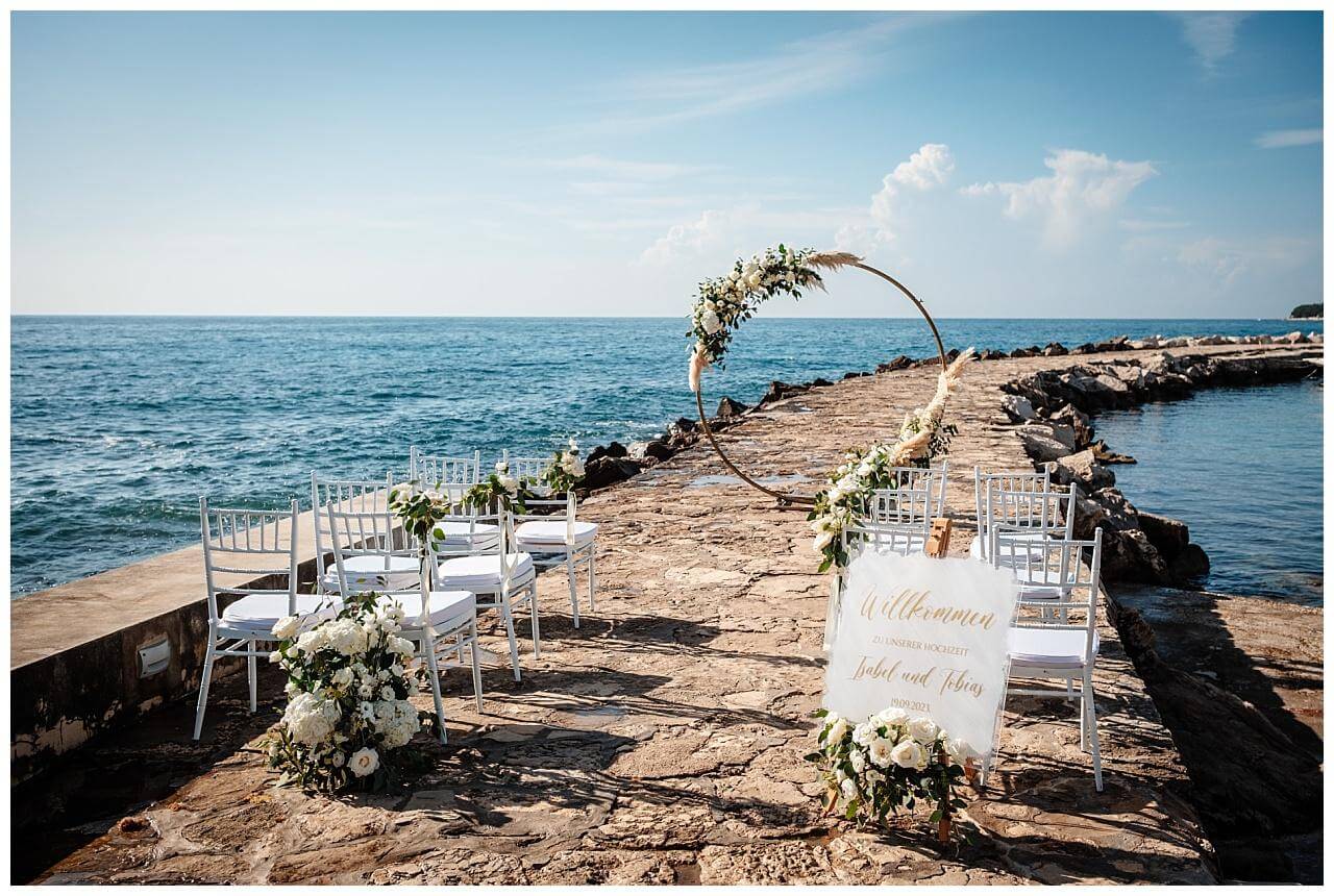 Traubogen aus Holz mit boho Blume Dekoration auf einen Steinweg im Meer und mit Glas Willkommensschild mit Goldener Schrift bei einer Hochzeit in Kroatien